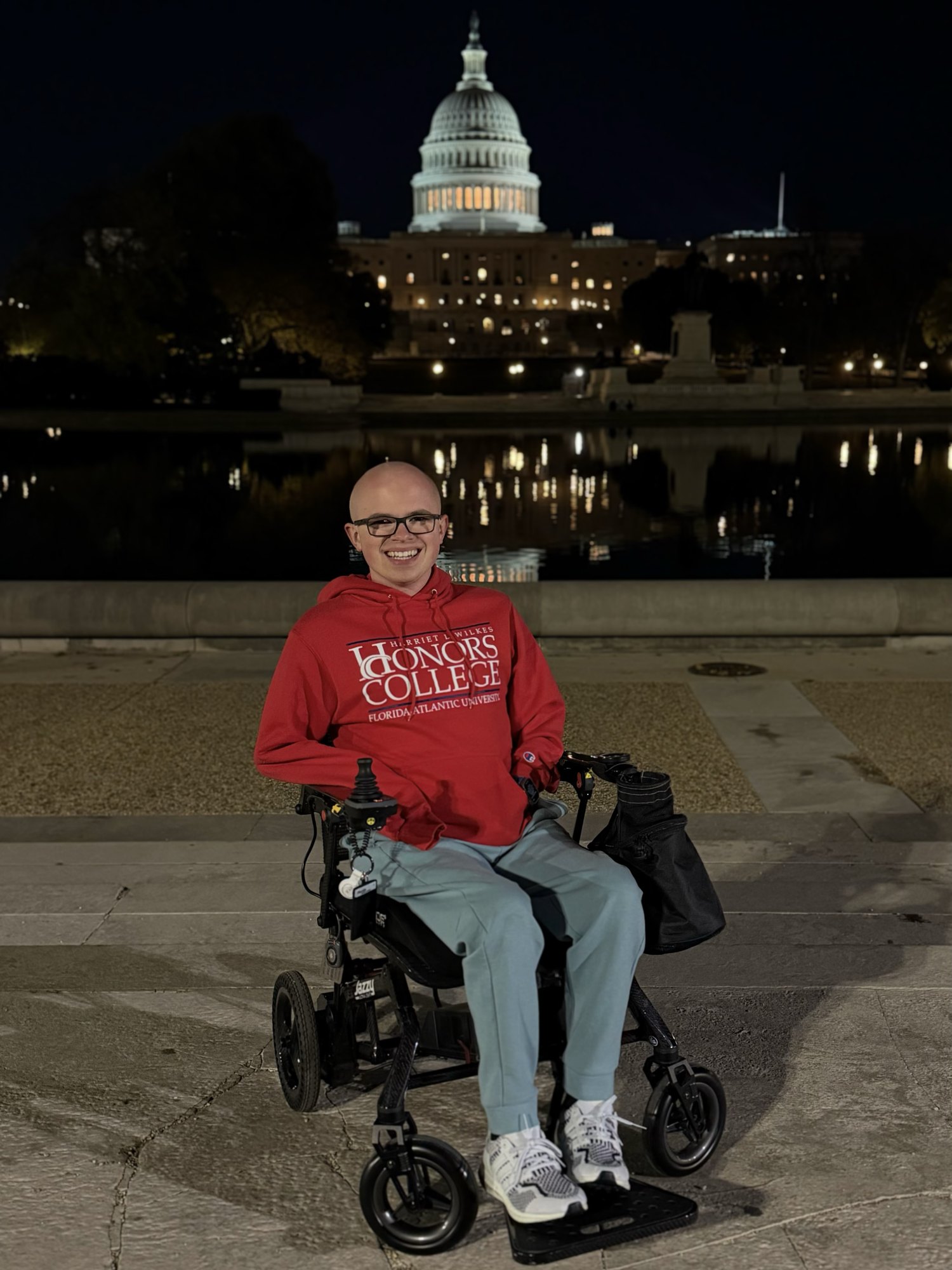 Ryleigh Newman sitting in his power wheelchair at night in front of the lit United States Capitol dome in Washington, D.C., wearing a red FAU Honors College hoodie.