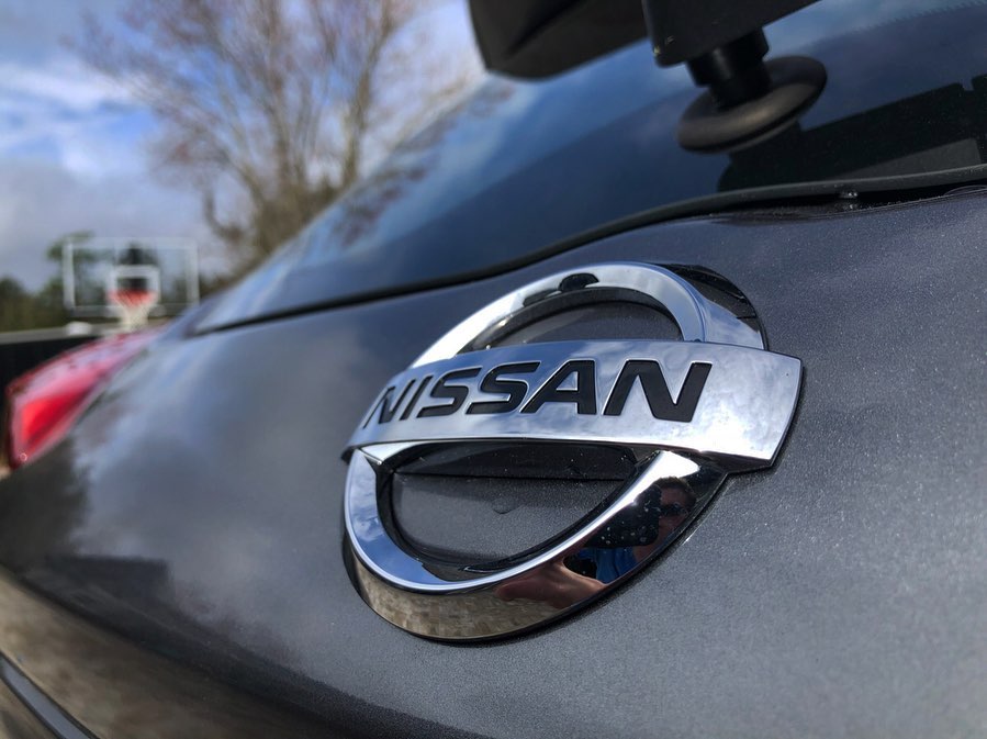 A close-up of the chrome Nissan emblem on the rear of the gray Kicks, reflecting a blue sky and bare tree branches.