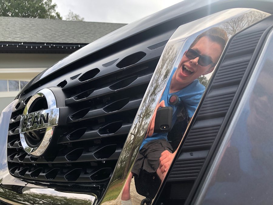 The front grille and chrome Nissan badge of the Kicks, with Ryleigh Newman laughing as he photographs himself reflected in the polished surface.