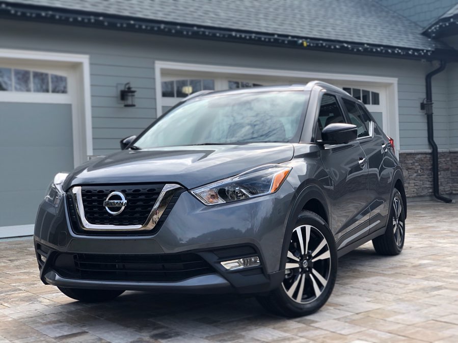 A gunmetal gray 2019 Nissan Kicks SR parked on a paver driveway in front of a pale green house, photographed from the front three-quarter angle on an overcast day.