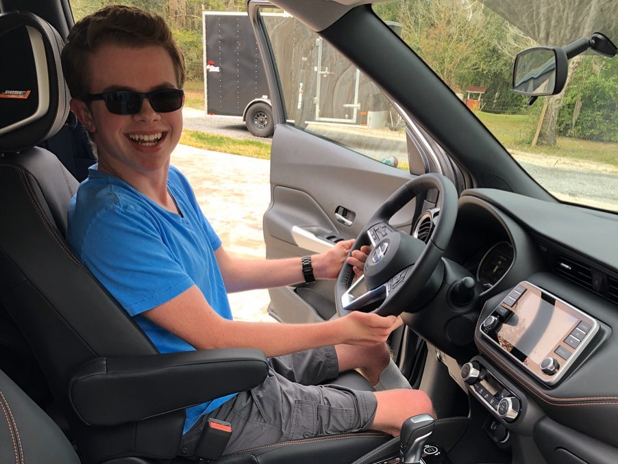 Ryleigh Newman sitting in the driver's seat of the Kicks in a blue T-shirt and sunglasses, smiling over his shoulder at the camera, with his hand resting near the wheel and a trailer and trees visible out the open passenger door.