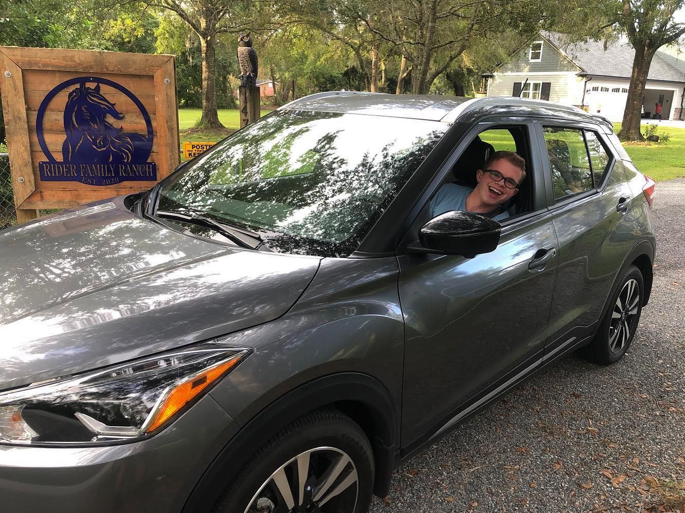 Ryleigh Newman behind the wheel of the gray Kicks with the driver's window down, leaning out and smiling, parked on a gravel drive beside a wooden Rider Family Ranch sign with a horse silhouette.