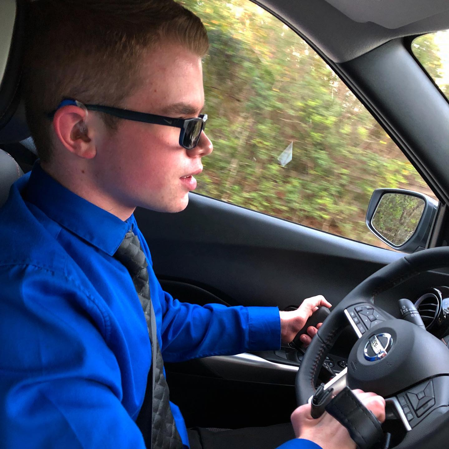 Ryleigh Newman in profile behind the wheel of his Nissan Kicks, wearing a blue dress shirt, tie, and sunglasses, one hand on the steering wheel and the other on the hand-control lever as trees blur past the window.