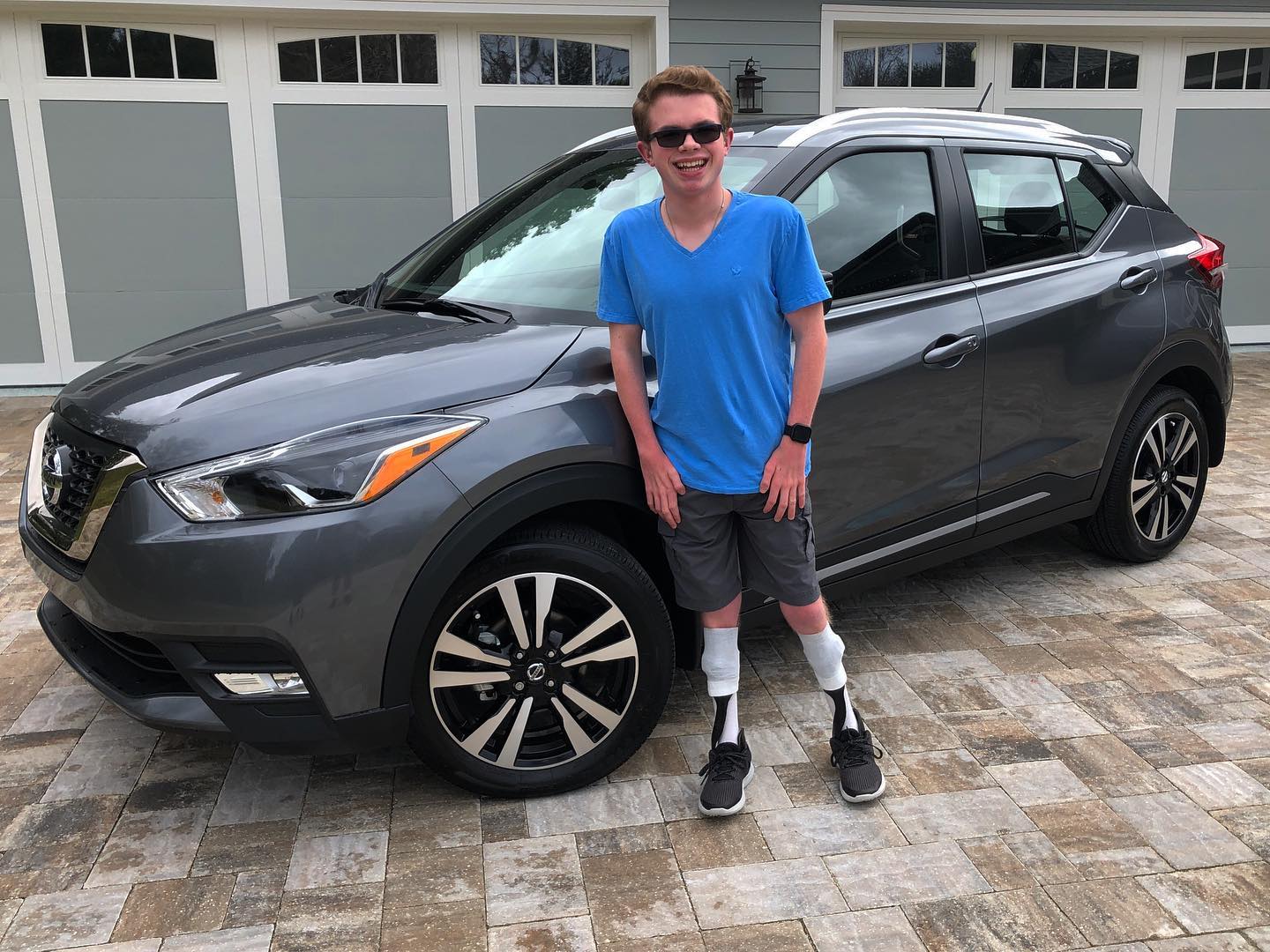 Ryleigh Newman standing in front of the gray Nissan Kicks in a blue T-shirt and shorts with leg braces visible, smiling on a paver driveway in front of garage doors.