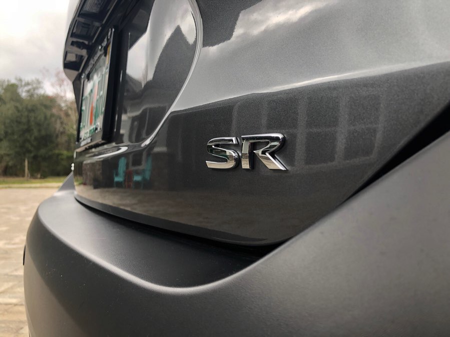 A close-up of the chrome SR trim badge on the rear of the gray Nissan Kicks, with a cloudy sky reflected in the paint.