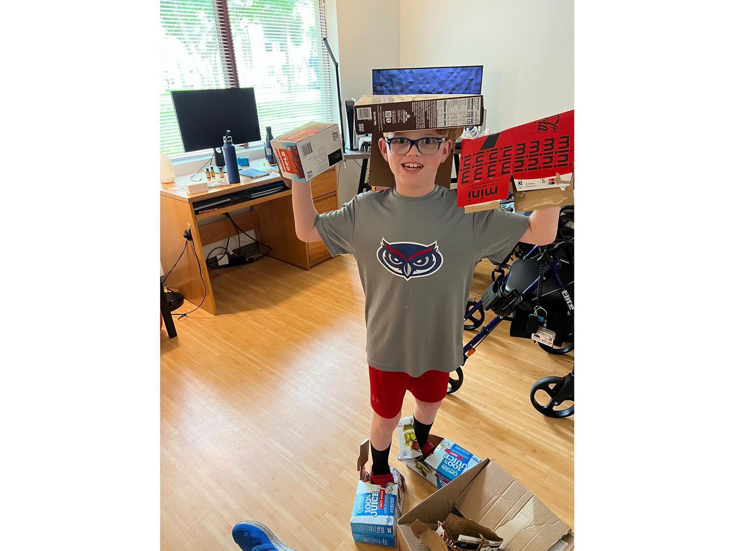 Ryleigh Newman's younger brother Emerson wearing an FAU owl shirt, playing with empty cardboard boxes stacked on his head and arms in the dorm room on move-in day.