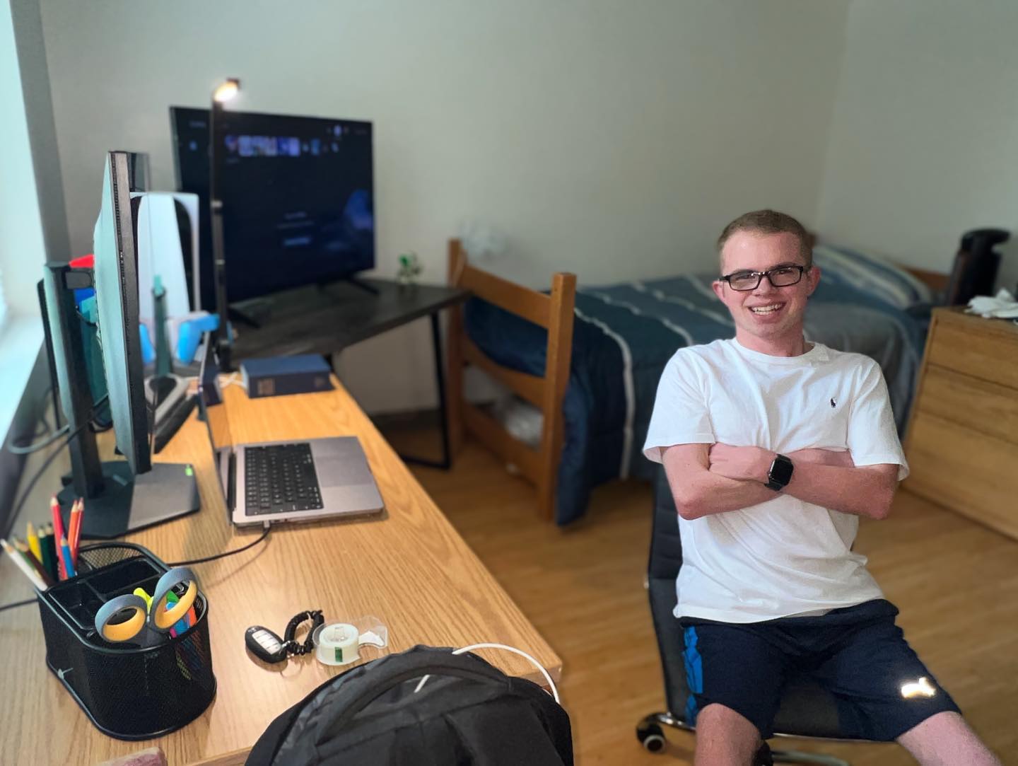 Ryleigh Newman sitting at his dorm desk on move-in day, arms crossed and smiling, with two monitors and a laptop set up on the wooden desk and a freshly made bed behind him.