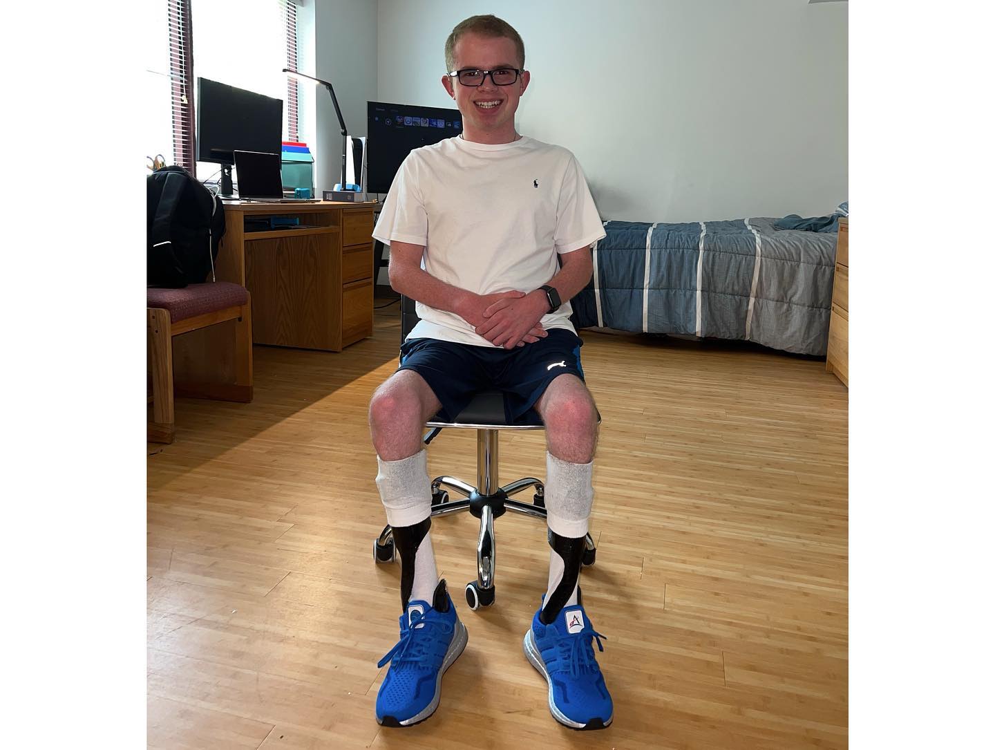 Ryleigh Newman sitting on a rolling stool in his dorm room wearing shorts, ankle-foot braces on both legs, and blue running shoes, smiling at the camera.