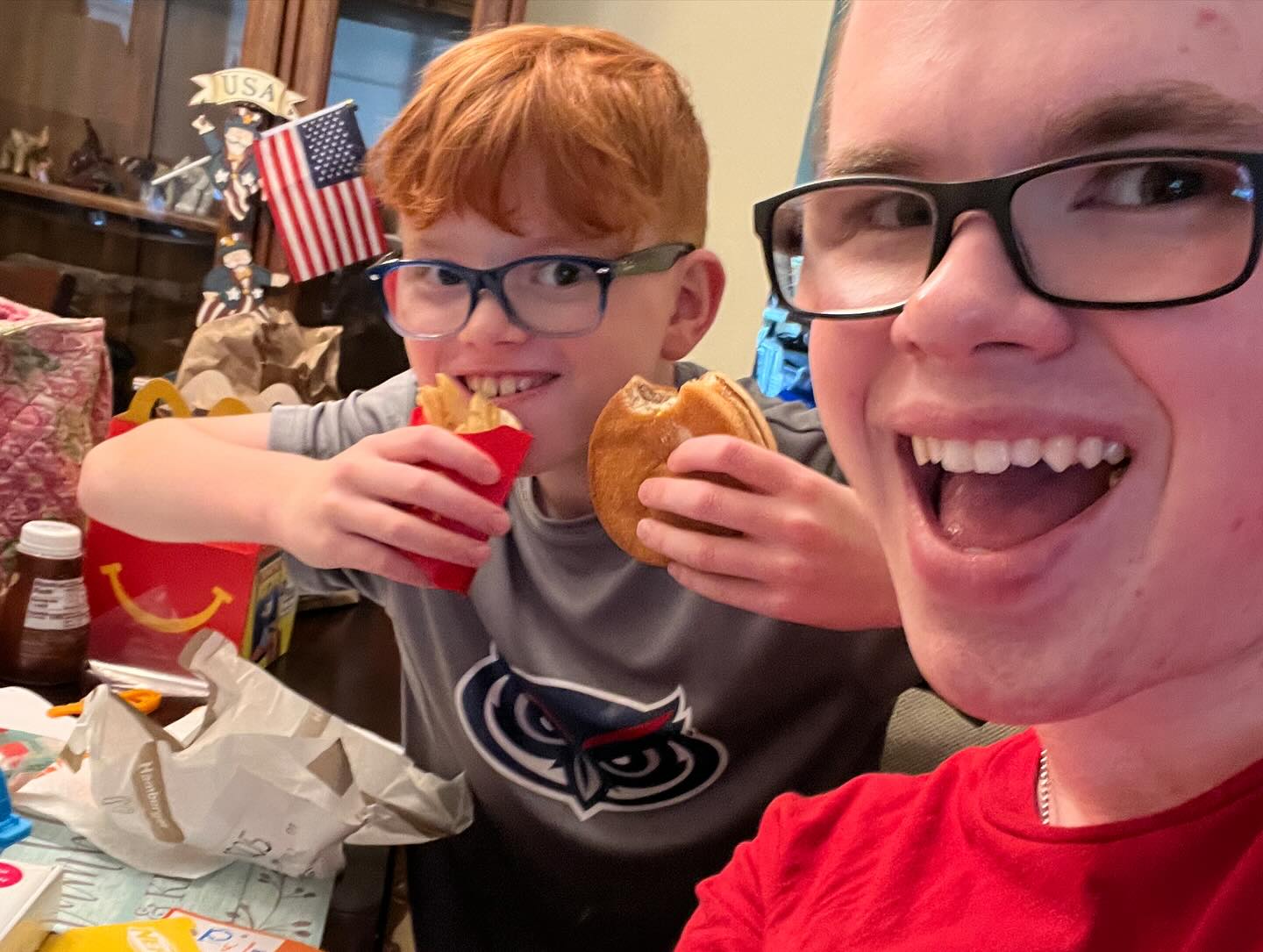 Ryleigh Newman in a red shirt sitting on the dorm floor unpacking snacks from a blue tote bag, smiling beside his younger brother Emerson, with a folded walker behind them.