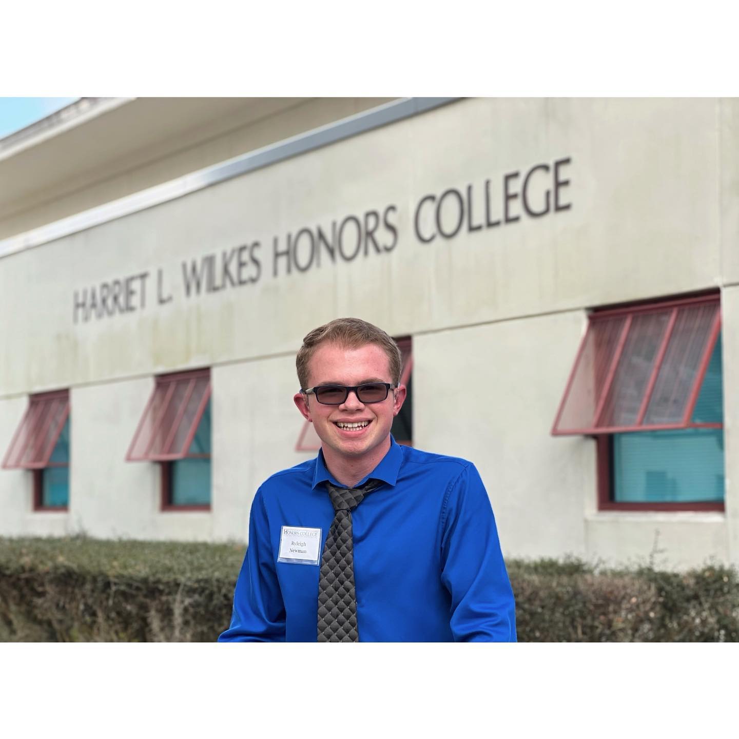 Ryleigh Newman in a blue dress shirt and tie with a Wilkes Honors College name tag, smiling in front of the building lettering that reads Harriet L. Wilkes Honors College.