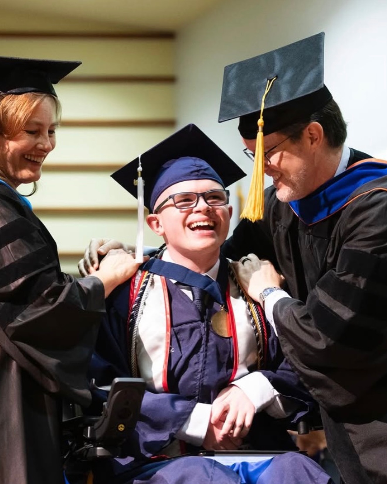 Two faculty members in academic regalia lean in to drape honor cords over a laughing Ryleigh Newman before the ceremony.