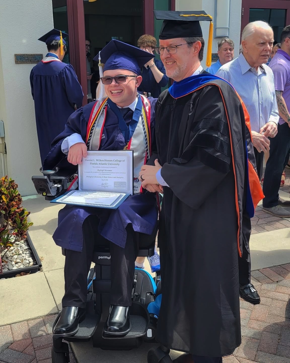 Ryleigh Newman shakes hands with a hooded professor in academic regalia just after receiving his diploma folder outside the venue.