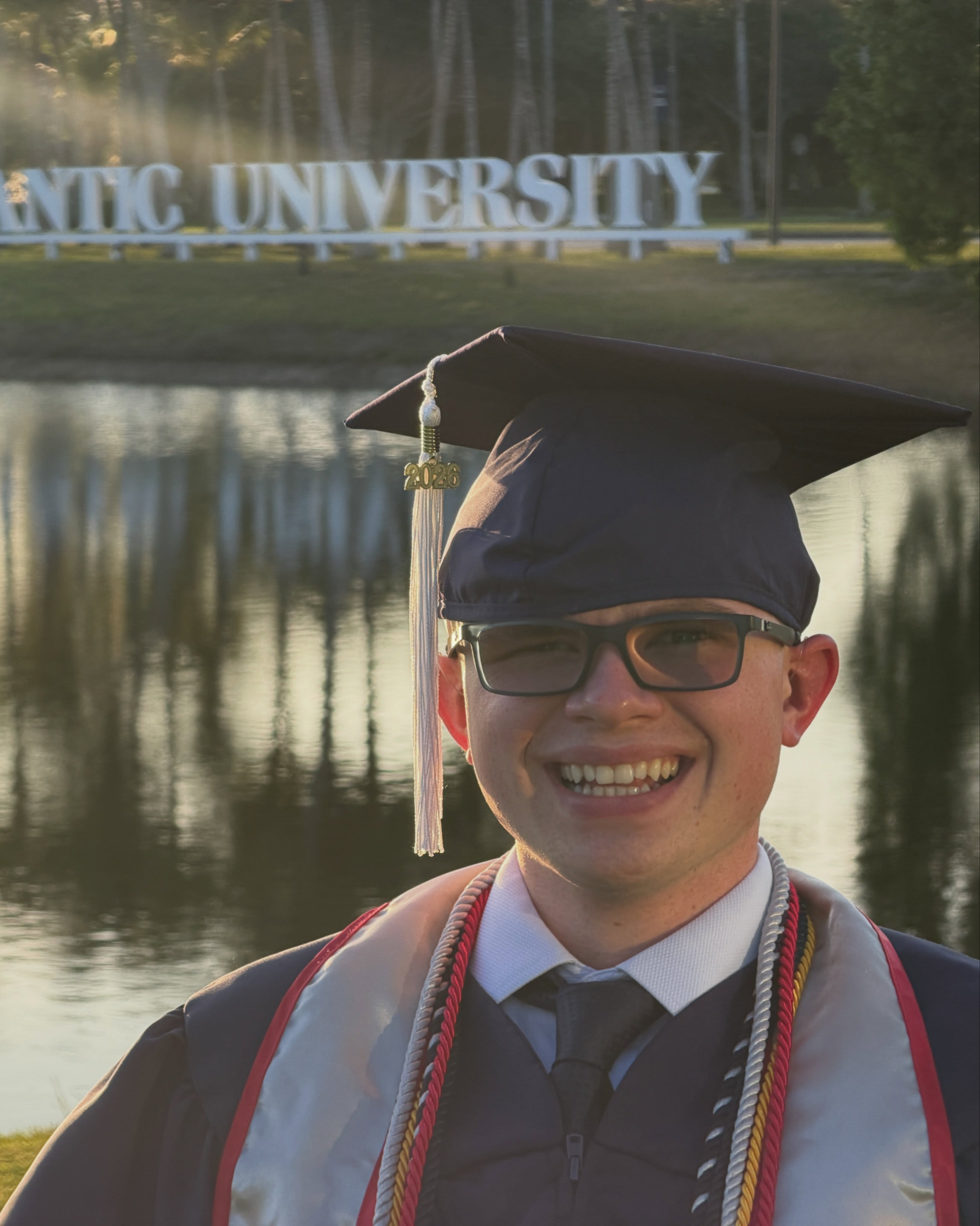 Close portrait of Ryleigh Newman in graduation cap with a 2026 tassel, the Florida Atlantic University lakeside sign blurred behind him.