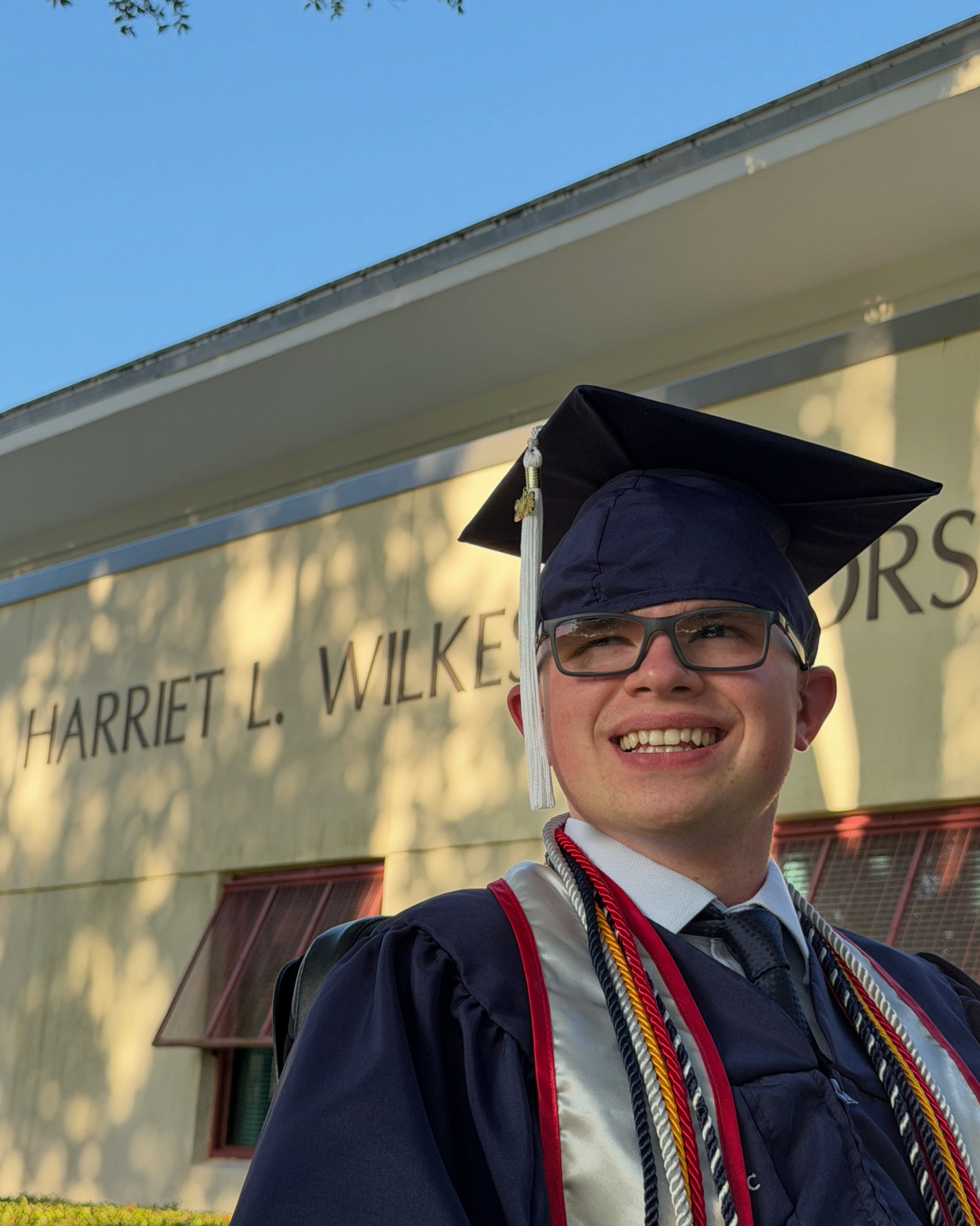 Ryleigh Newman in cap, gown, and honor cords in front of the Harriet L. Wilkes Honors College building.