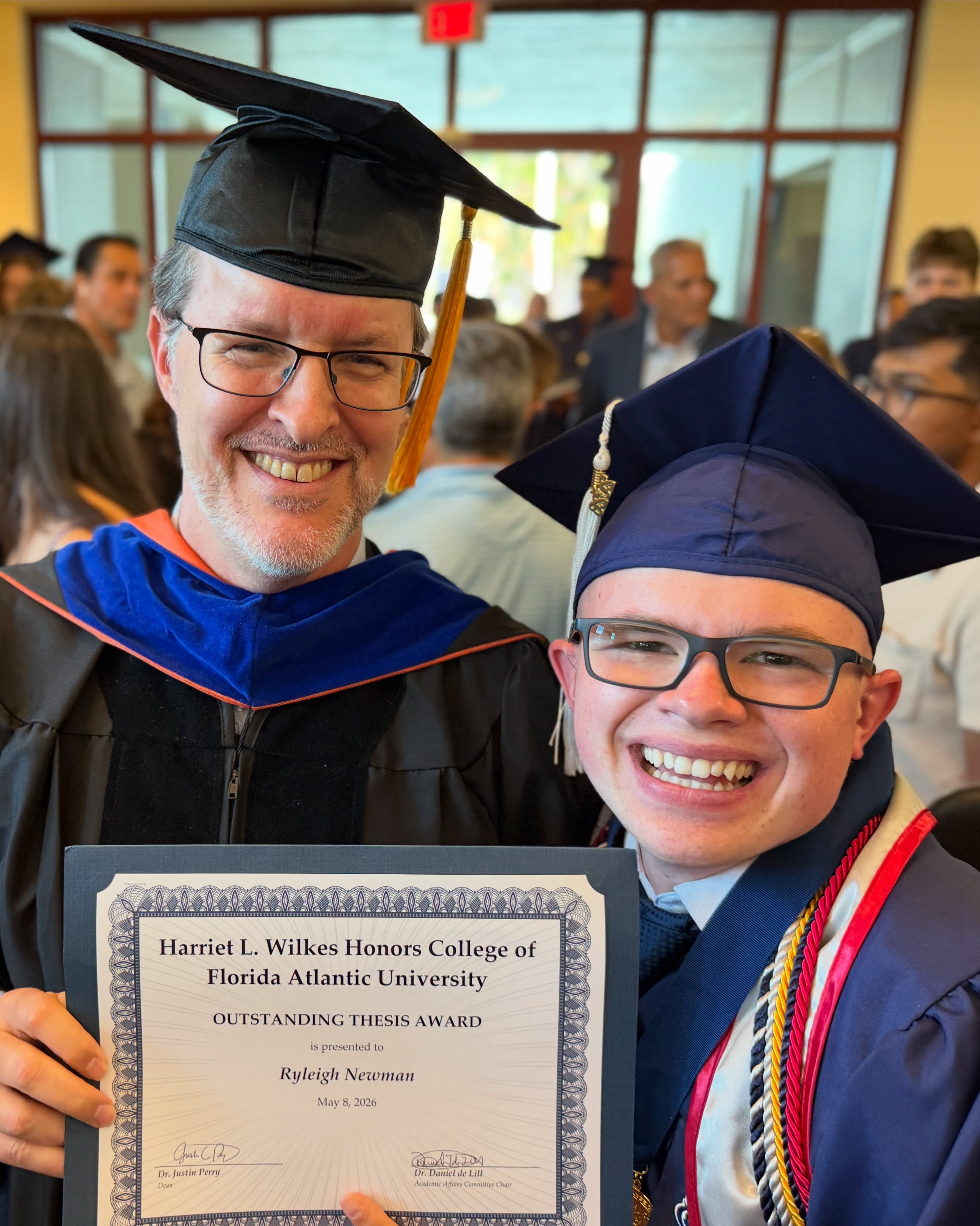 Ryleigh Newman and his thesis advisor, both in academic regalia, smiling next to the framed Outstanding Thesis Award certificate from the Harriet L. Wilkes Honors College.