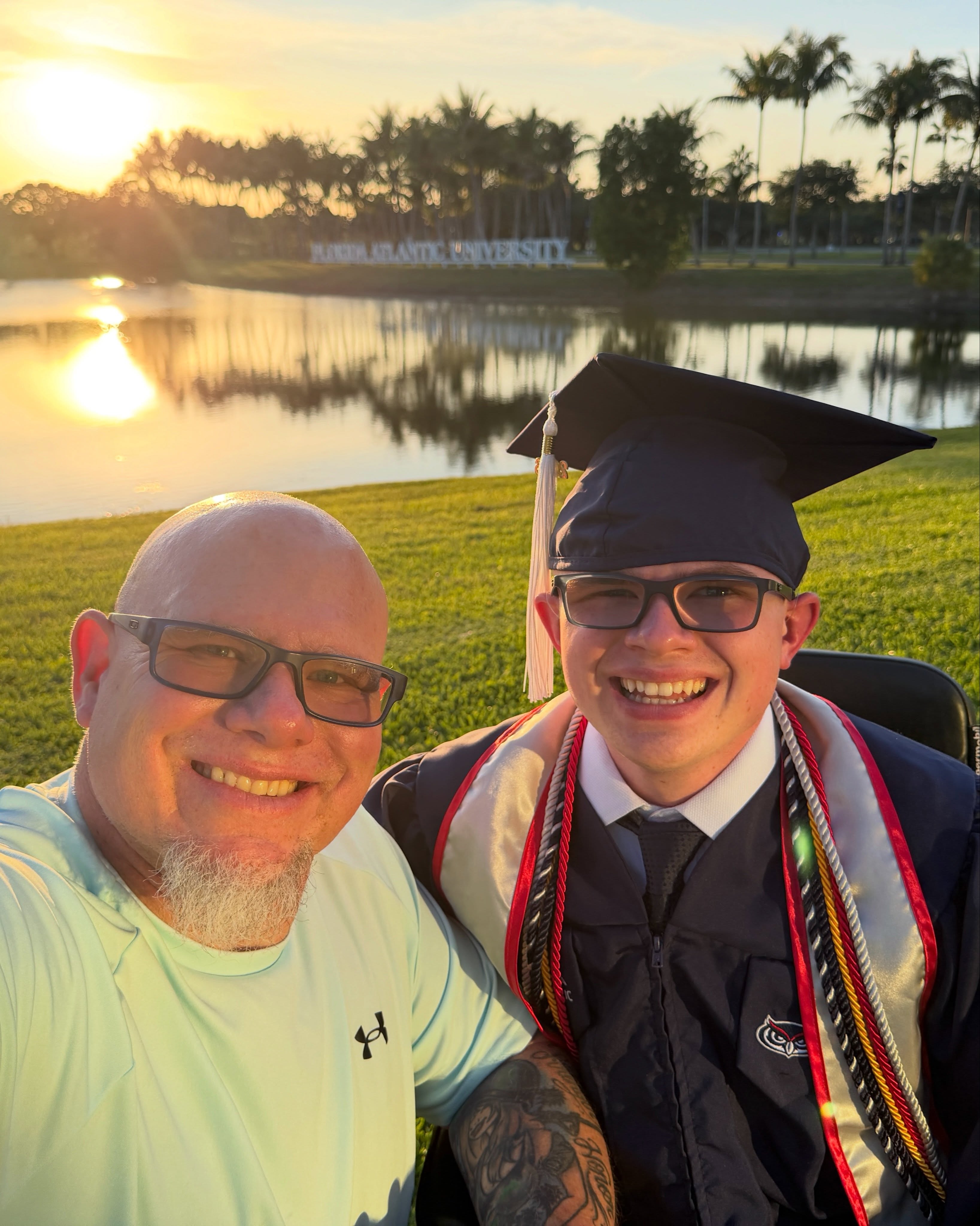 Ryleigh Newman in cap and honor cords and his father smiling together at sunset beside the Florida Atlantic University lake.