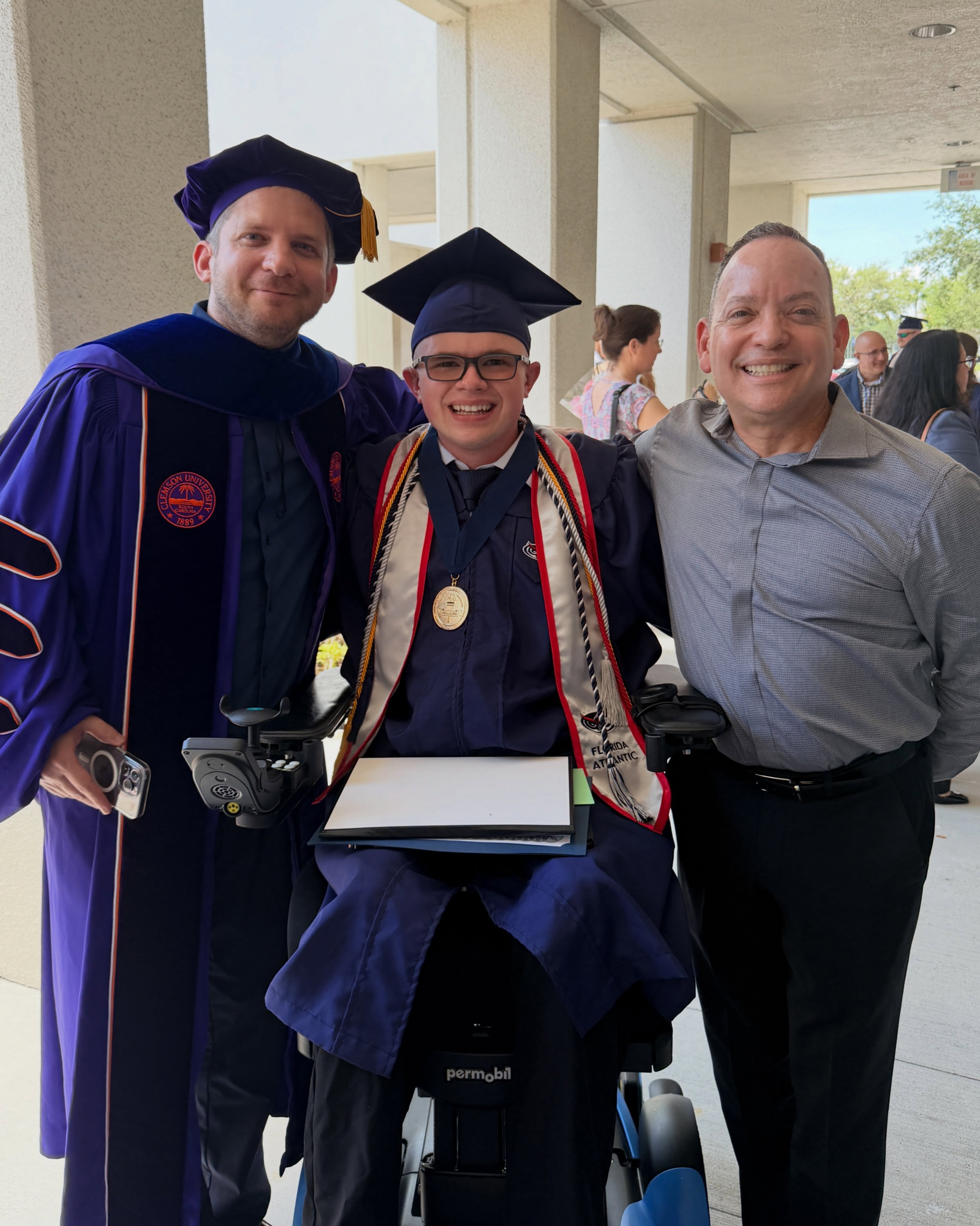 Ryleigh Newman in cap and gown holding his diploma and medallion, flanked by two faculty members in academic regalia outside the venue after the ceremony.