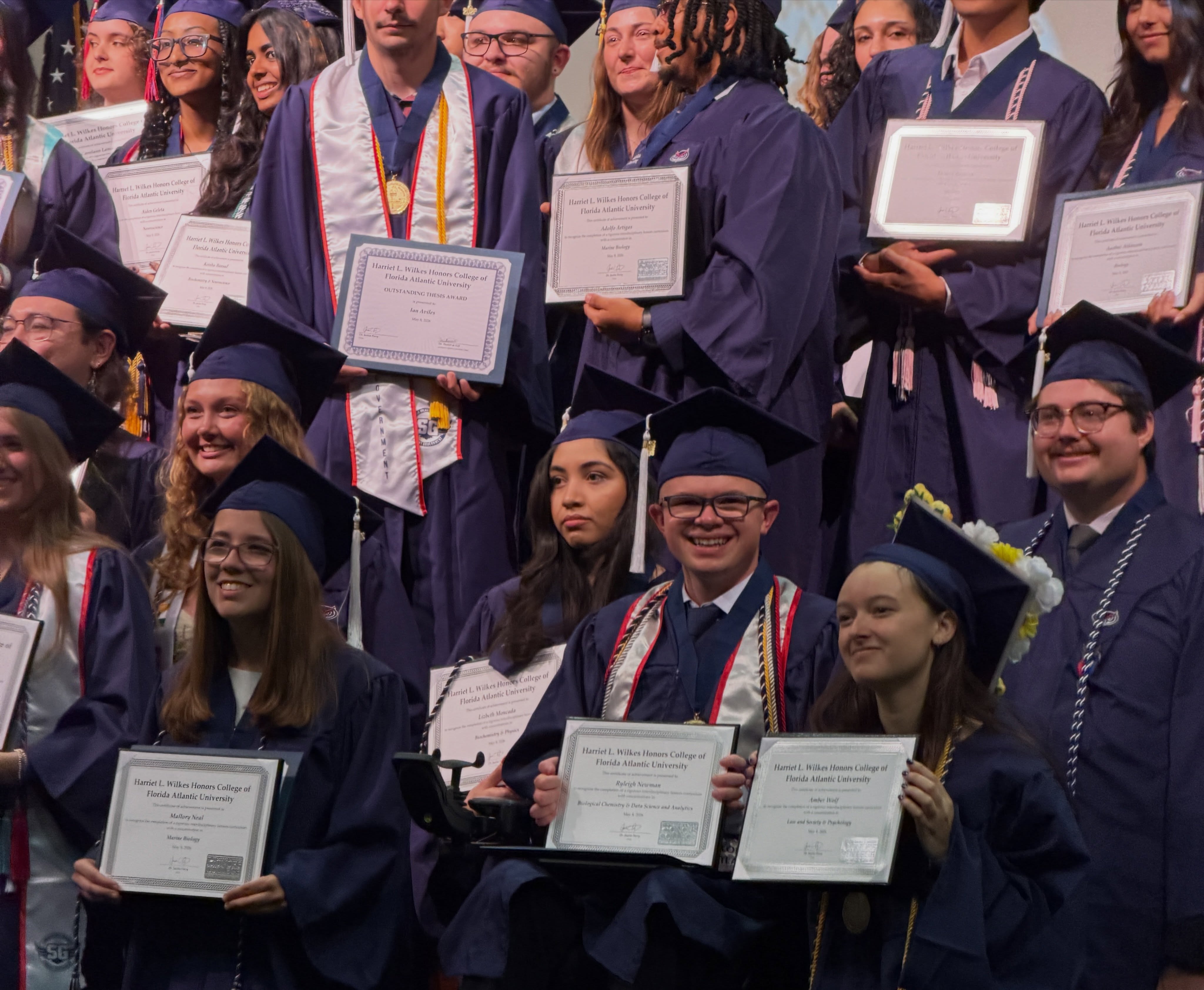 Ryleigh Newman smiling among a tight group of Wilkes Honors College graduates holding up their framed certificates after the ceremony.