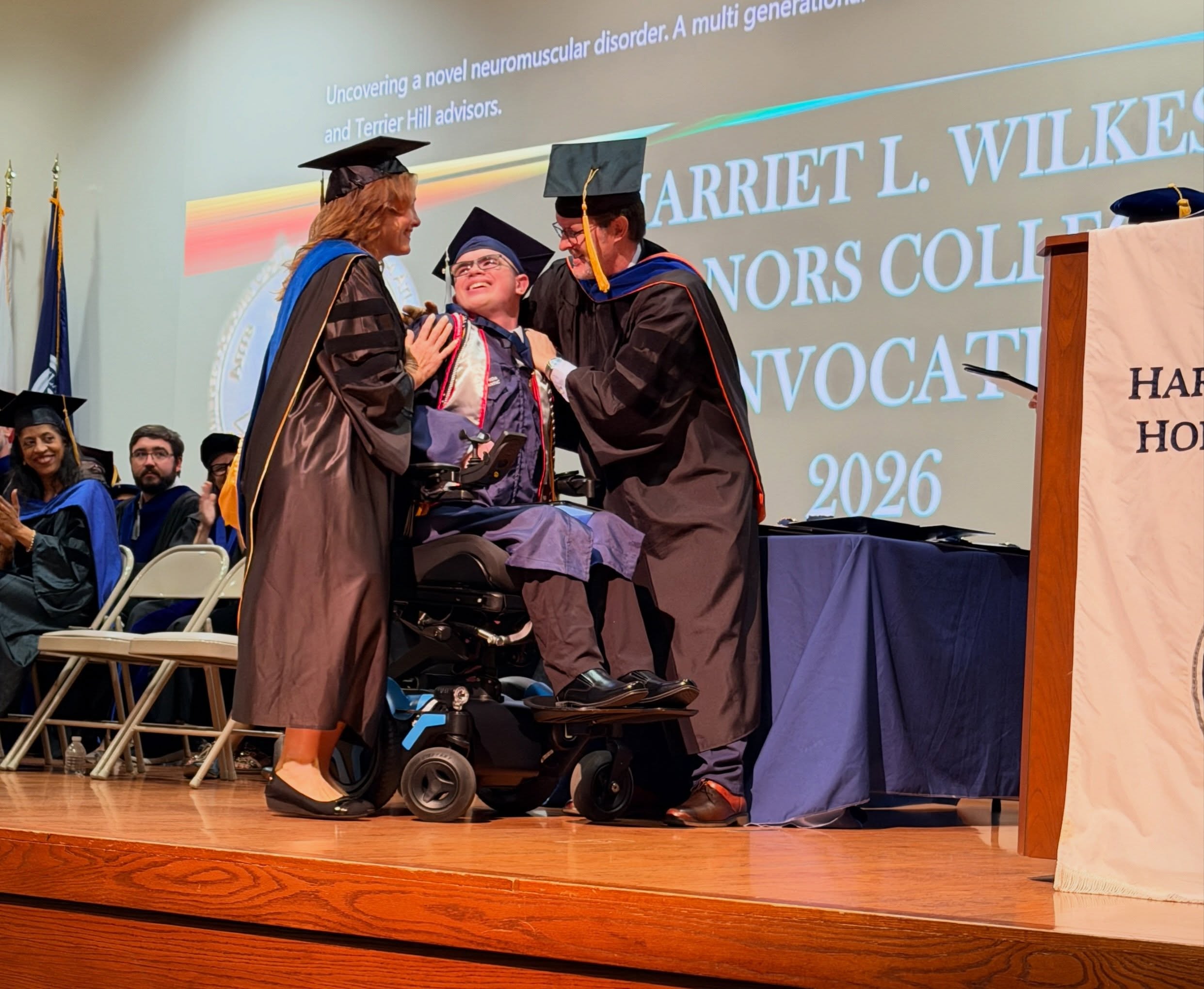 Ryleigh Newman laughing in his wheelchair on stage as two Wilkes Honors College faculty members in regalia lean in to congratulate him, with the 2026 Convocation slide projected behind them.
