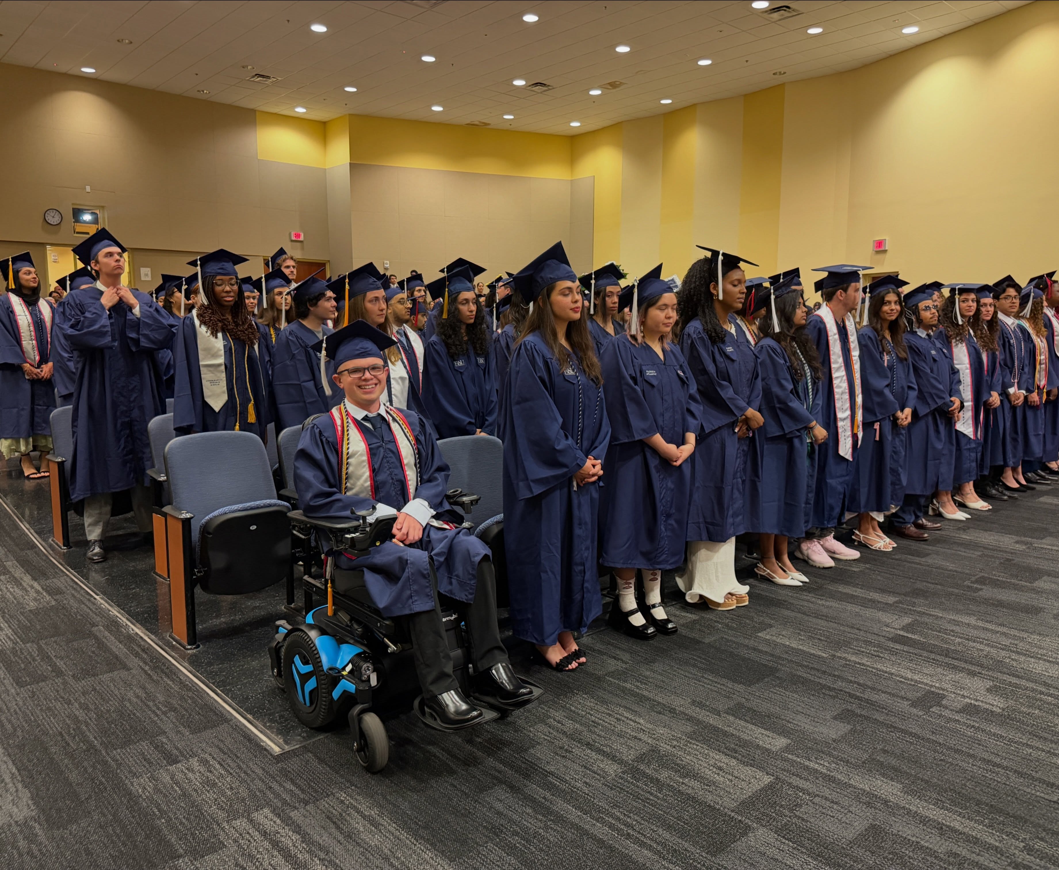 Ryleigh Newman in navy cap and gown waiting in his blue power wheelchair at the front of a long line of Wilkes Honors College graduates during the processional.