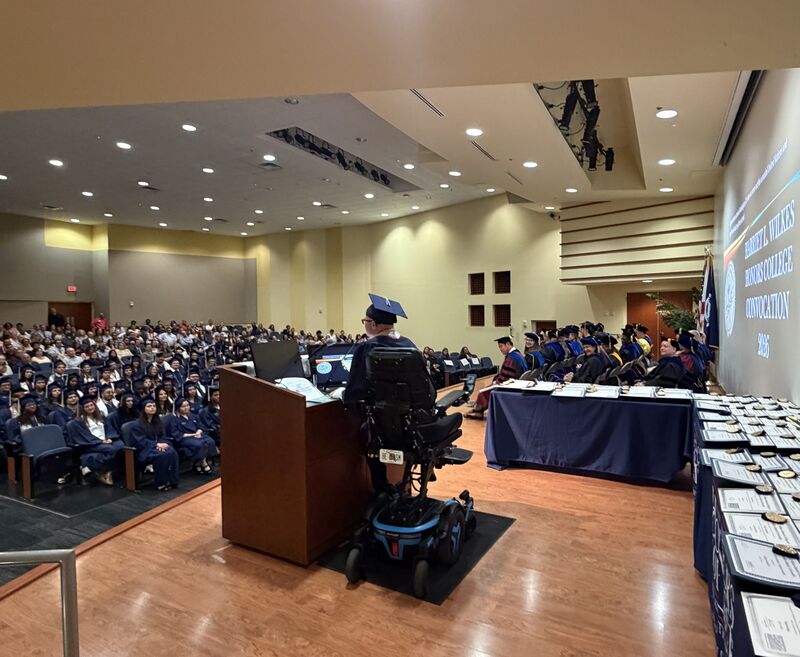Ryleigh Newman in cap and gown addressing the Wilkes Honors College Convocation from a podium in his power wheelchair.