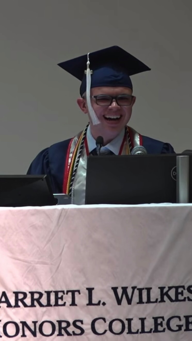 Ryleigh Newman in navy cap and gown laughing mid-sentence at the podium, with the Harriet L. Wilkes Honors College banner draped in front of him.