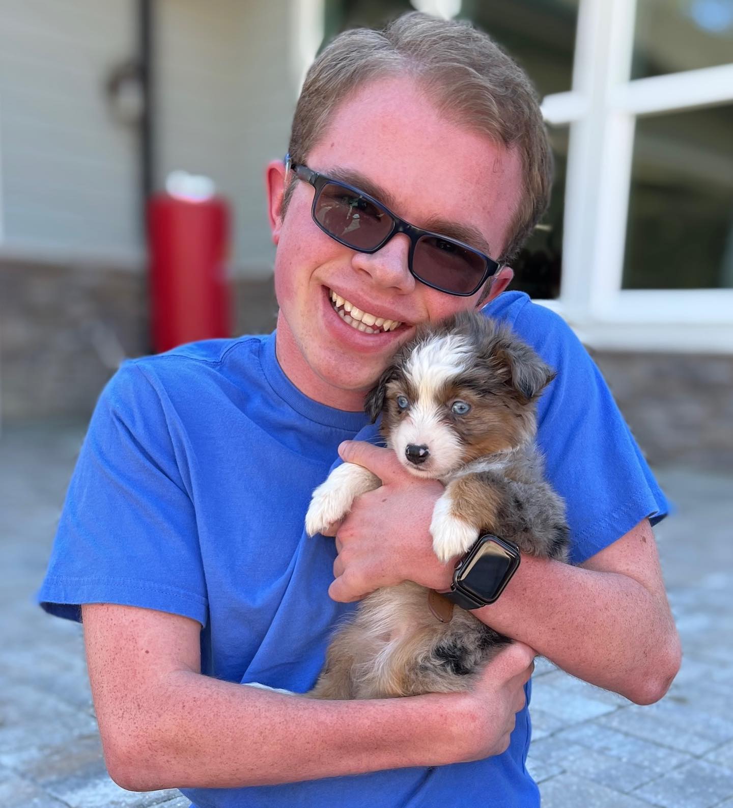 Ryleigh Newman in sunglasses and a blue shirt smiling broadly while holding a fluffy blue merle mini Aussie puppy with one piercing blue eye against his chest.