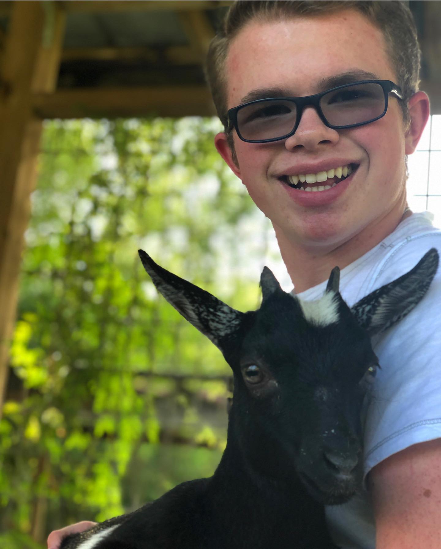 Ryleigh Newman smiling under a shelter while cradling a young black goat with white-tipped ears against his shoulder.
