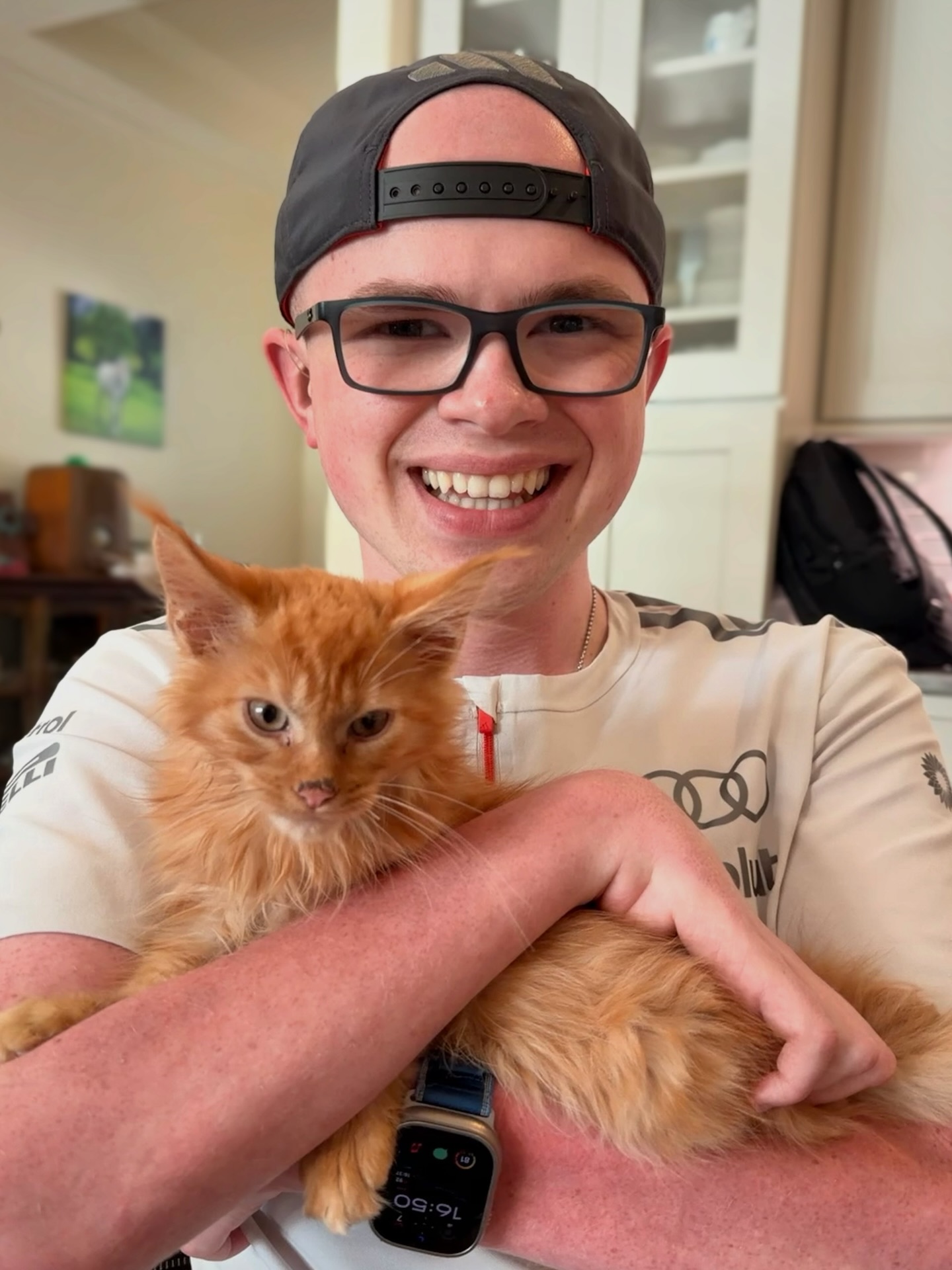 Ryleigh Newman in a backward cap and glasses smiling while holding a fluffy orange Maine Coon kitten cradled in his arms indoors.