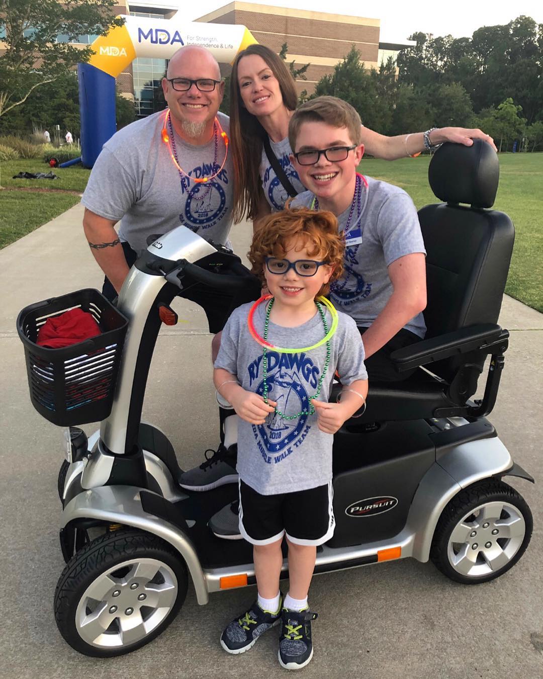 Ryleigh Newman with his family on his mobility scooter at the 2019 MDA Muscle Walk, all in matching team shirts under the MDA arch.