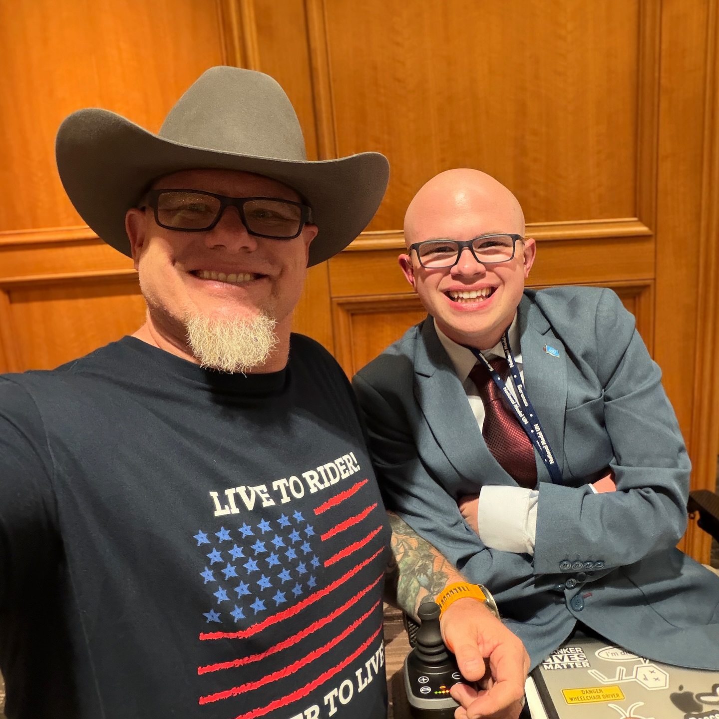 Ryleigh Newman in a blue blazer smiling beside his dad, who is wearing a cowboy hat and a 'Live to Ride' flag T-shirt, at the Japan conference in Washington.
