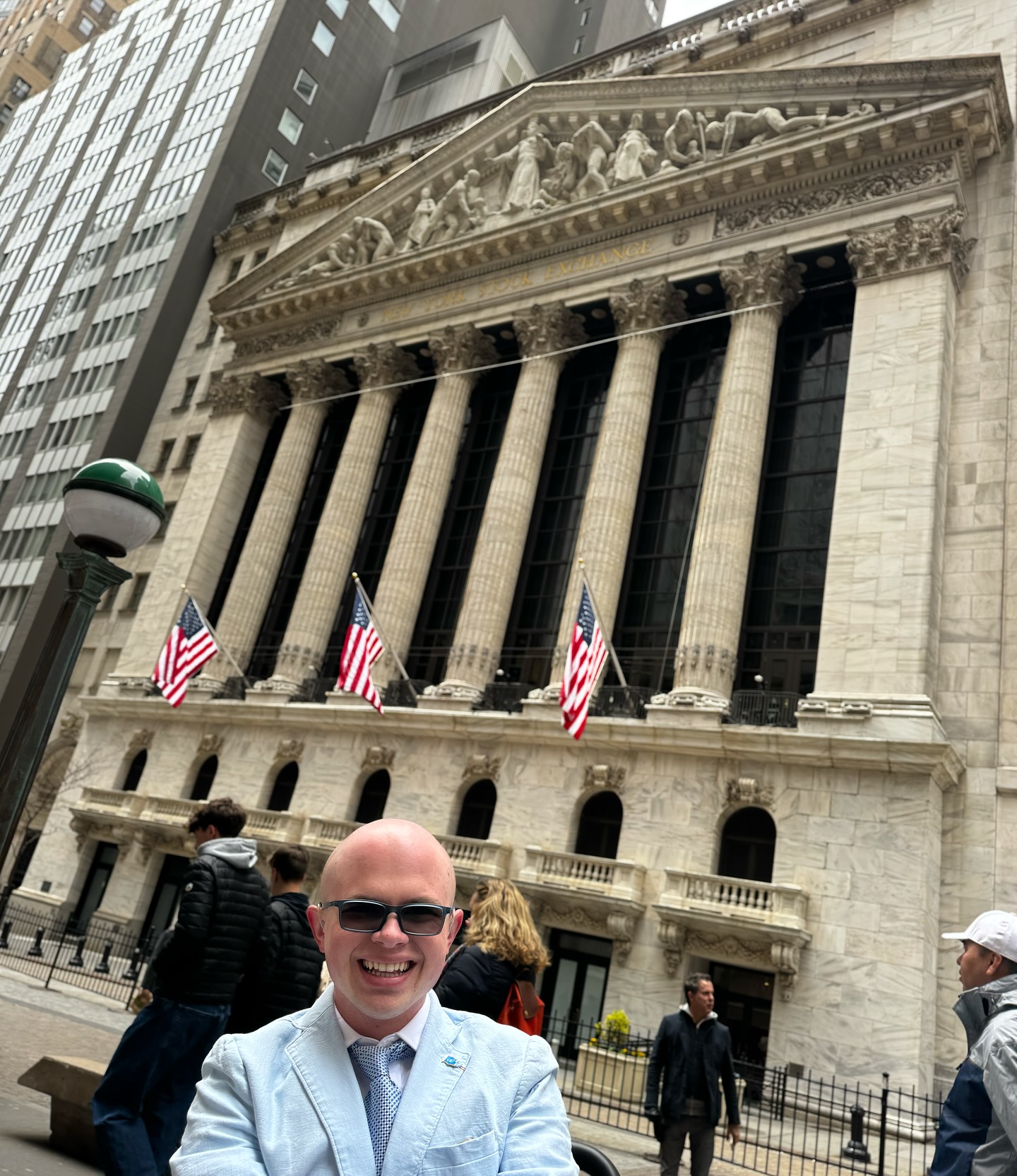The columned facade of the New York Stock Exchange with American flags, Ryleigh Newman in front in sunglasses.