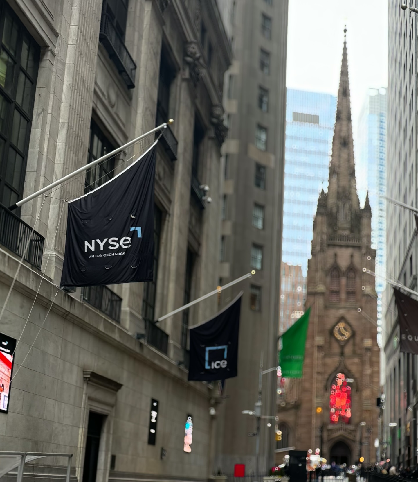 NYSE and ICE banners hanging on the exchange building with the Gothic spire of Trinity Church framed at the end of Wall Street.