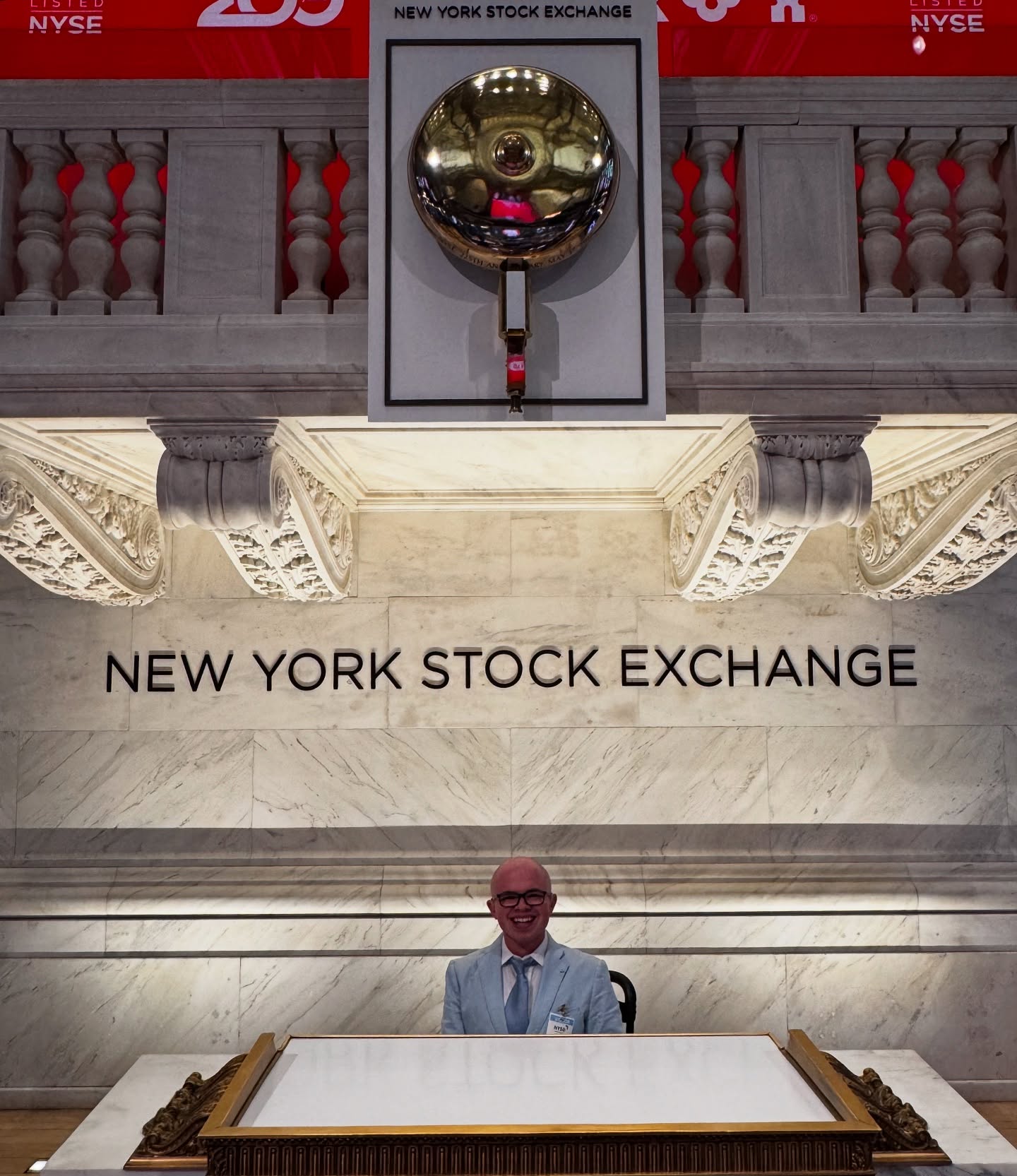 Ryleigh Newman smiling on the trading floor of the New York Stock Exchange, with the curved 'New York Stock Exchange' desk, a 200 Years sign, and walls of market screens behind him.