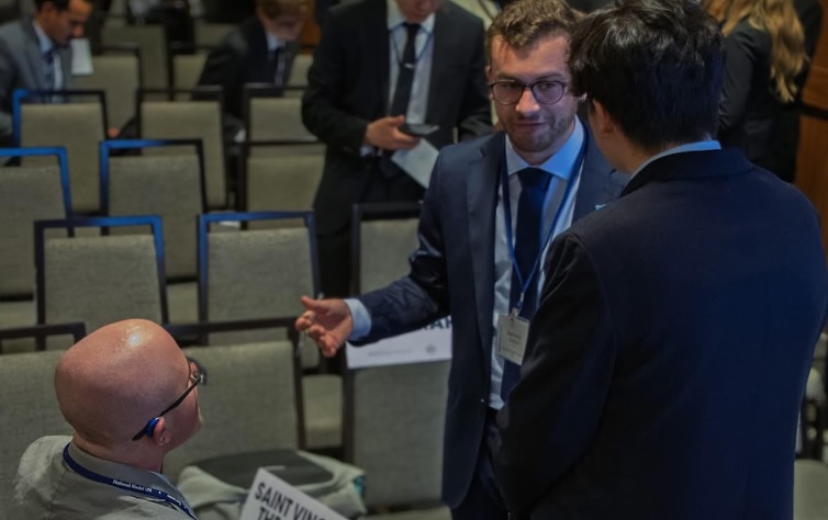 Ryleigh Newman, seen from behind with his Saint Vincent placard, talking with two standing delegates among rows of empty blue chairs during a committee break.