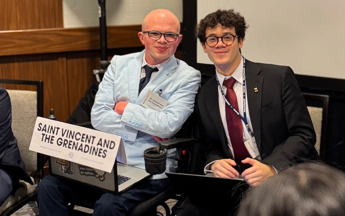 Ryleigh Newman in a light blue blazer seated beside a fellow delegate in a wood-paneled committee room, with a Saint Vincent and the Grenadines placard on the table in front of him.