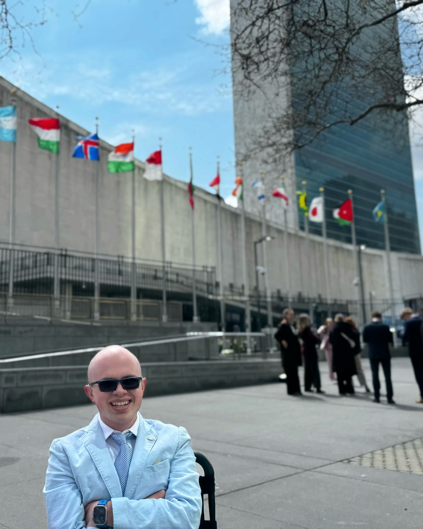 Ryleigh Newman in a light blue blazer and sunglasses in front of United Nations Headquarters, with the row of member-state flags and the Secretariat tower behind him under a blue sky.