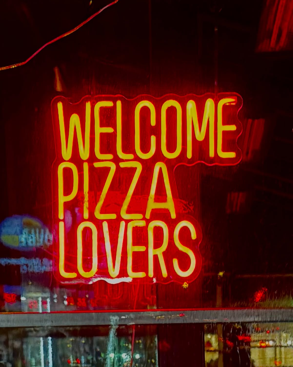 A red and yellow neon sign in a window reading 'Welcome Pizza Lovers.'