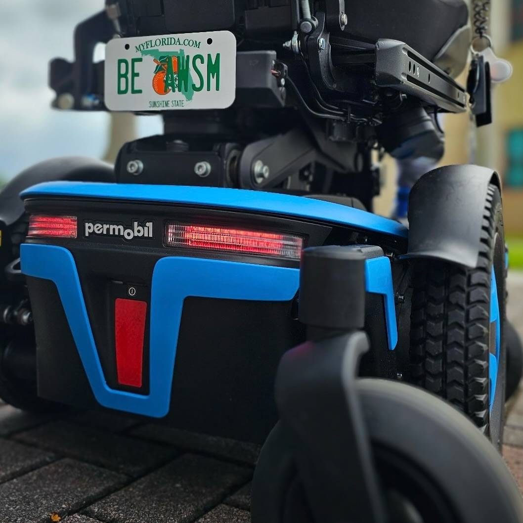 Close-up of the rear of the blue Permobil M3 Corpus showing the illuminated red light bar with the Permobil logo, a red reflector on the blue accent panel, the treaded drive wheel, and a Florida BE AWSM license plate above.