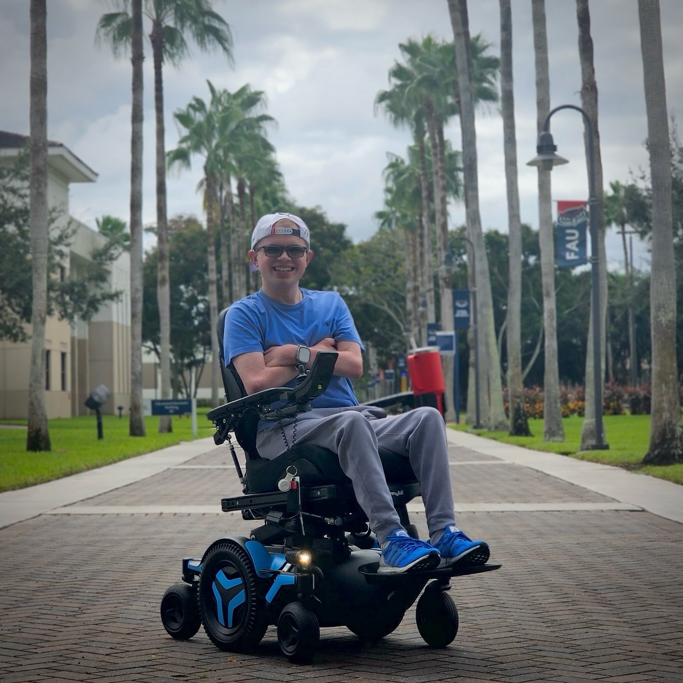 Ryleigh Newman in his Permobil M3 Corpus power wheelchair on a palm-lined campus path at FAU.