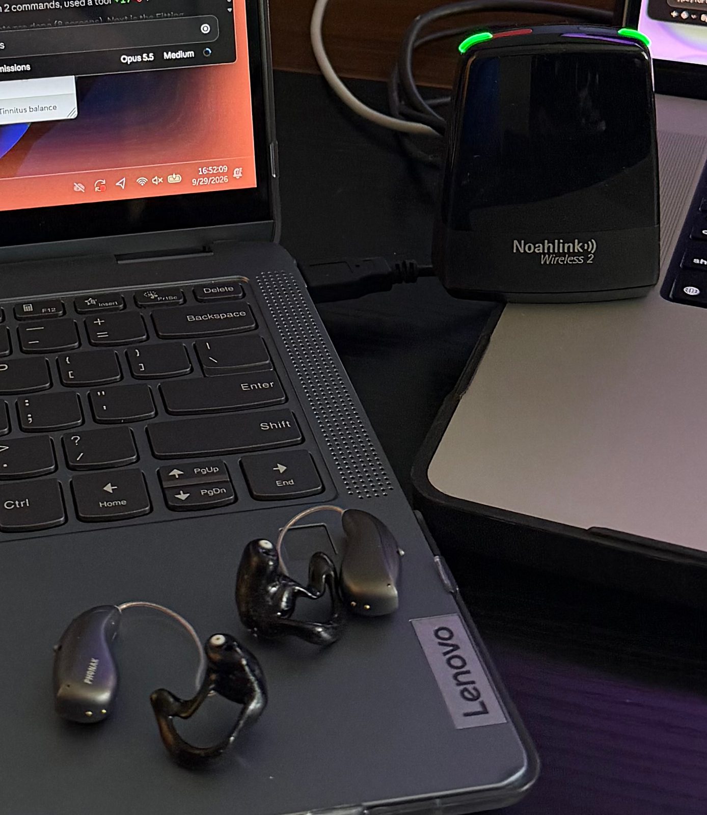 Close-up of two Phonak Audéo Sphere receiver-in-canal hearing aids with glossy black custom shells lying on a dark gray laptop, with the black Noahlink Wireless 2 programming dongle behind them, its status lights glowing green.