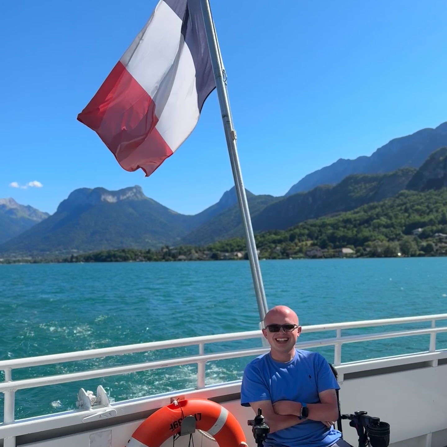 The turquoise water of Lake Annecy seen from a boat railing, with a sharply peaked green mountain rising across the lake under a clear blue sky.