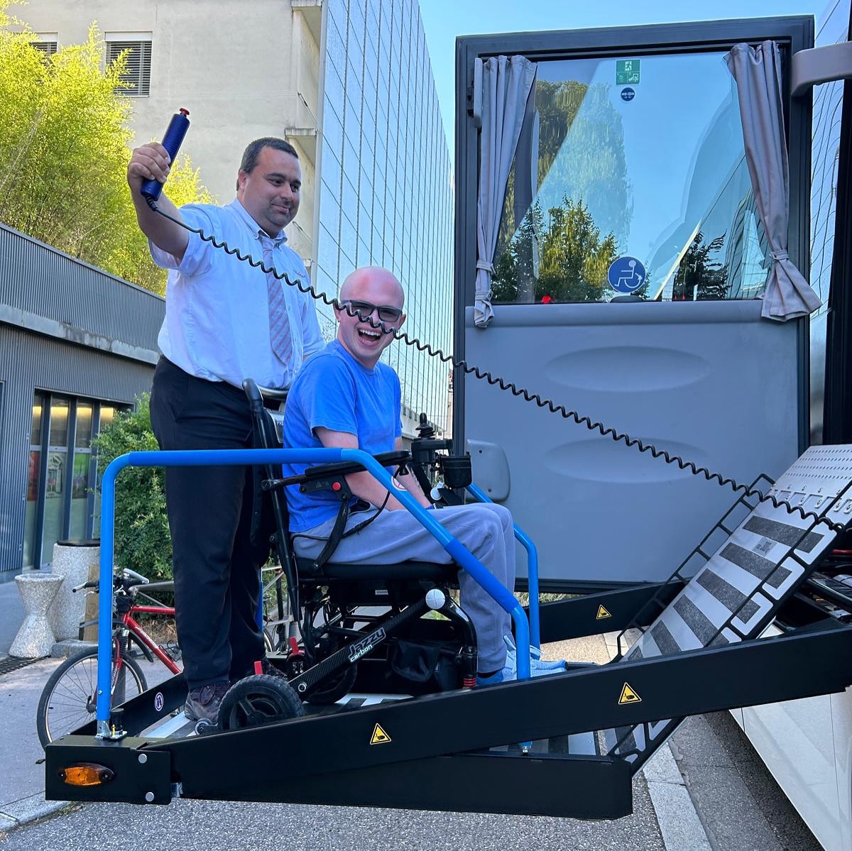 Ryleigh Newman in his wheelchair on a bridge in Annecy old town, with the medieval stone Palais de l'Isle and a flower-lined canal behind him under a bright sky.