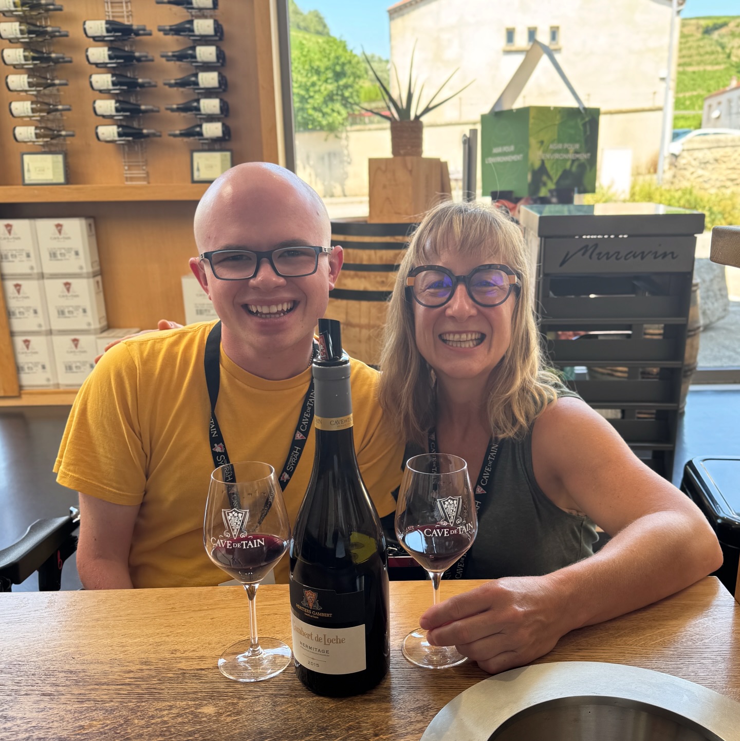 Ryleigh Newman and his mother smiling side by side at the Cave de Tain tasting bar, with two glasses of red wine and a bottle in front of them and a wall of wine behind.