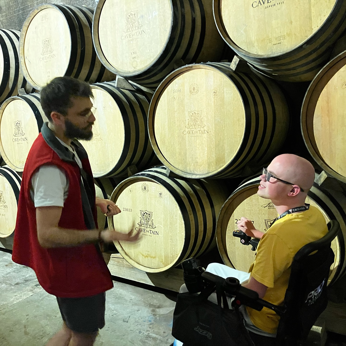 Ryleigh Newman in his wheelchair looking up at a host in a red vest who is explaining the oak barrels stacked floor to ceiling in the Cave de Tain cellar.