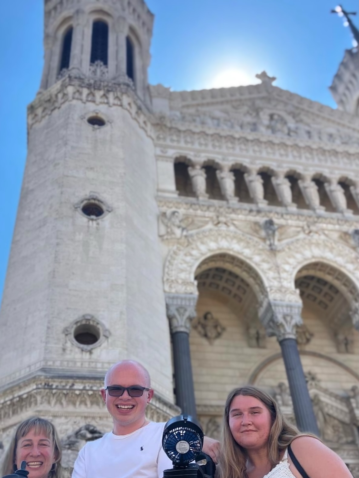 Ryleigh Newman and others smiling below the towering carved white stone facade of the Basilica of Notre-Dame de Fourviere in Lyon, with the sun cresting the rooftop and a clear blue sky.