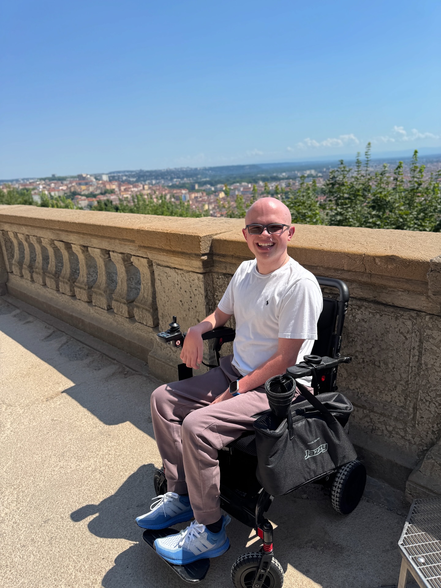 Ryleigh Newman in his power wheelchair on the terrace below the Fourviere basilica, with the rooftops of Lyon spread out behind him under a bright blue sky.