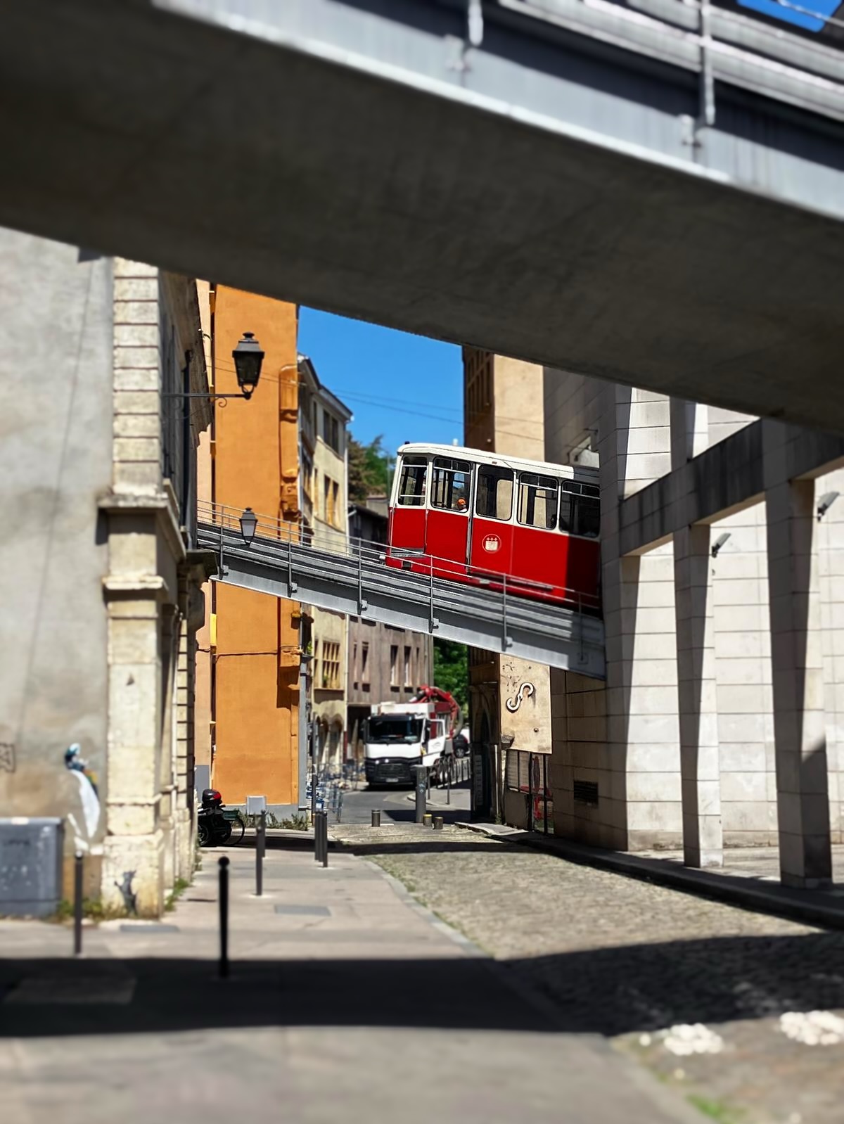 A bright red Lyon funicular tram crossing an elevated track between old ochre and cream buildings, framed by a concrete overpass, on a sunny day.