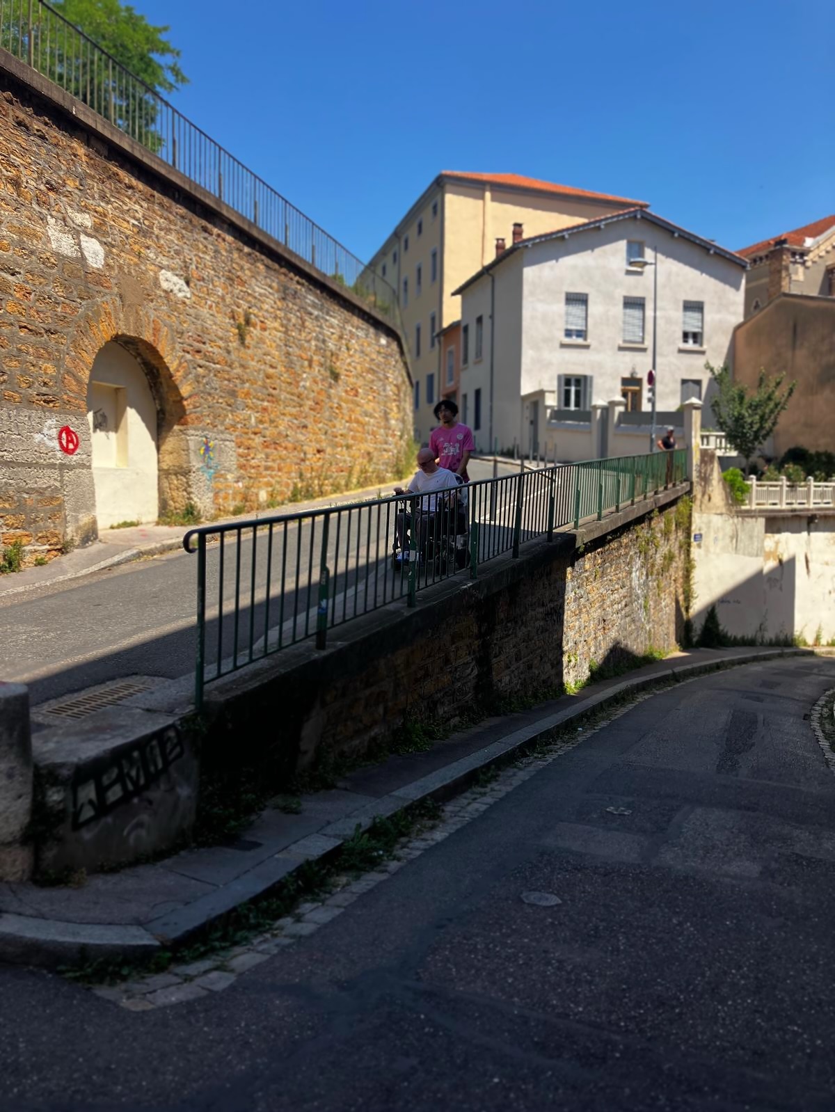 Ryleigh Newman in his power wheelchair on a narrow walkway above an old stone street in Lyon, with a weathered arched wall and pastel buildings climbing the hill behind him.