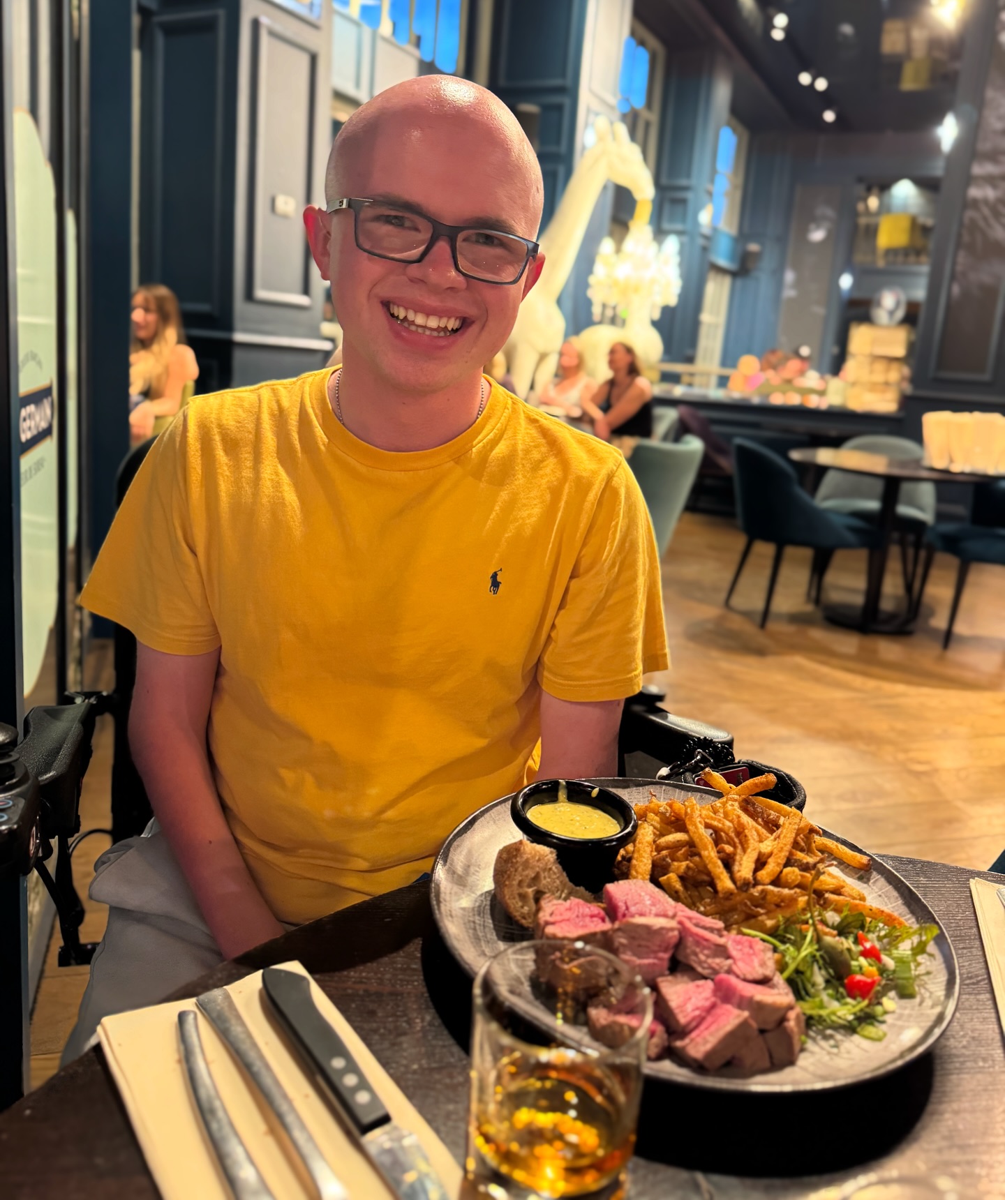 Ryleigh Newman smiling at a restaurant table over a plate of sliced steak, golden frites, and a sauce pot, with a giraffe statue glowing in the dining room behind him.